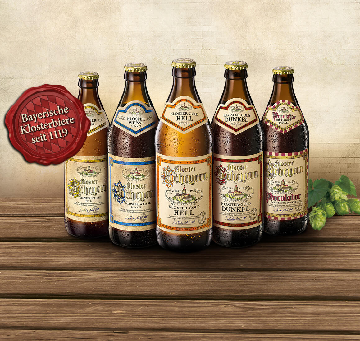 Fünf Flaschen Bier stehen auf einem Holztisch vor einer Wand.